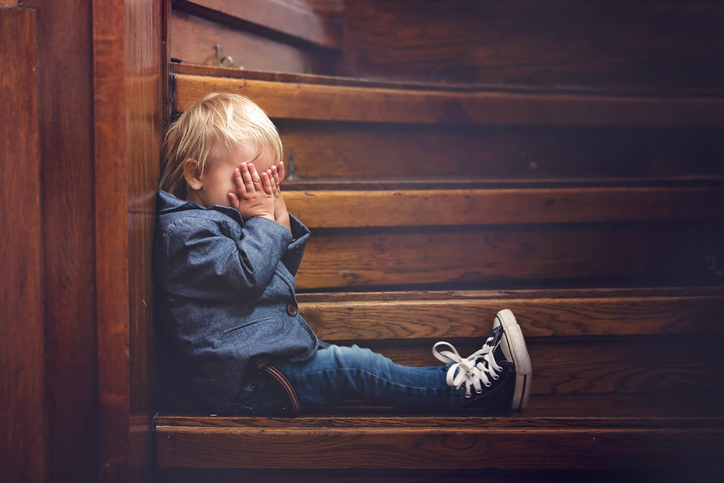 A young child sitting alone on wooden stairs, covering their face with their hands to represent the emotional impact and isolation often addressed in childhood trauma therapy.
